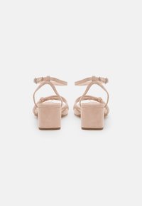 Beige suede heeled sandals with ankle straps, open back, and block heels. Features knotted upper straps and a smooth texture.