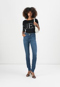 Top nero con logo metallico, jeans blu a vita alta e tacchi neri con cinturini. Una borsa bianca è tenuta nella mano sinistra.