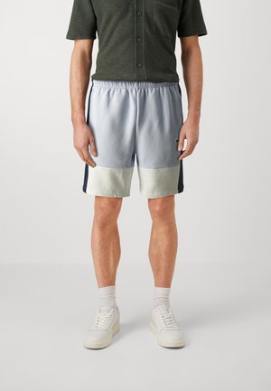 Man draagt lichtblauwe en beige color-blocked korte broek, donkergroene korte mouwen blouse met knopen, witte sokken en witte sneakers, staand.