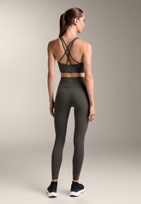 Temno zelena športna modrček s prepletenimi naramnicami, v kombinaciji z visokimi temno zelenimi leggins in črnimi športnimi čevlji. Gladka tekstura.