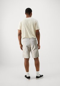Tricou din bumbac bej și pantaloni scurți gri cu buzunare, purtați cu șosete albe și pantofi sport negri, văzuți din spate pe un fundal simplu.