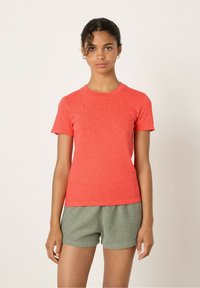 Kurzärmliges korallenfarbiges T-Shirt aus einem weichen, strukturierten Stoff. Verfügt über einen runden Hals und eine lässige Passform. Getragen mit grünen Shorts.