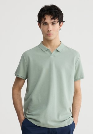 Polo shirt verde pallido con maniche corte, design a scollo a V, texture a coste e dettagli a pattern discreti. Indossato con pantaloni blu scuro.
