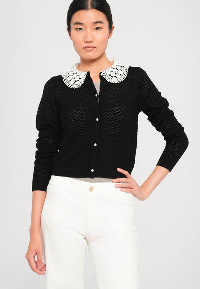 Black, knit cardigan with a white lace collar, featuring a button front and long sleeves. Paired with white pants with heart-shaped pockets.