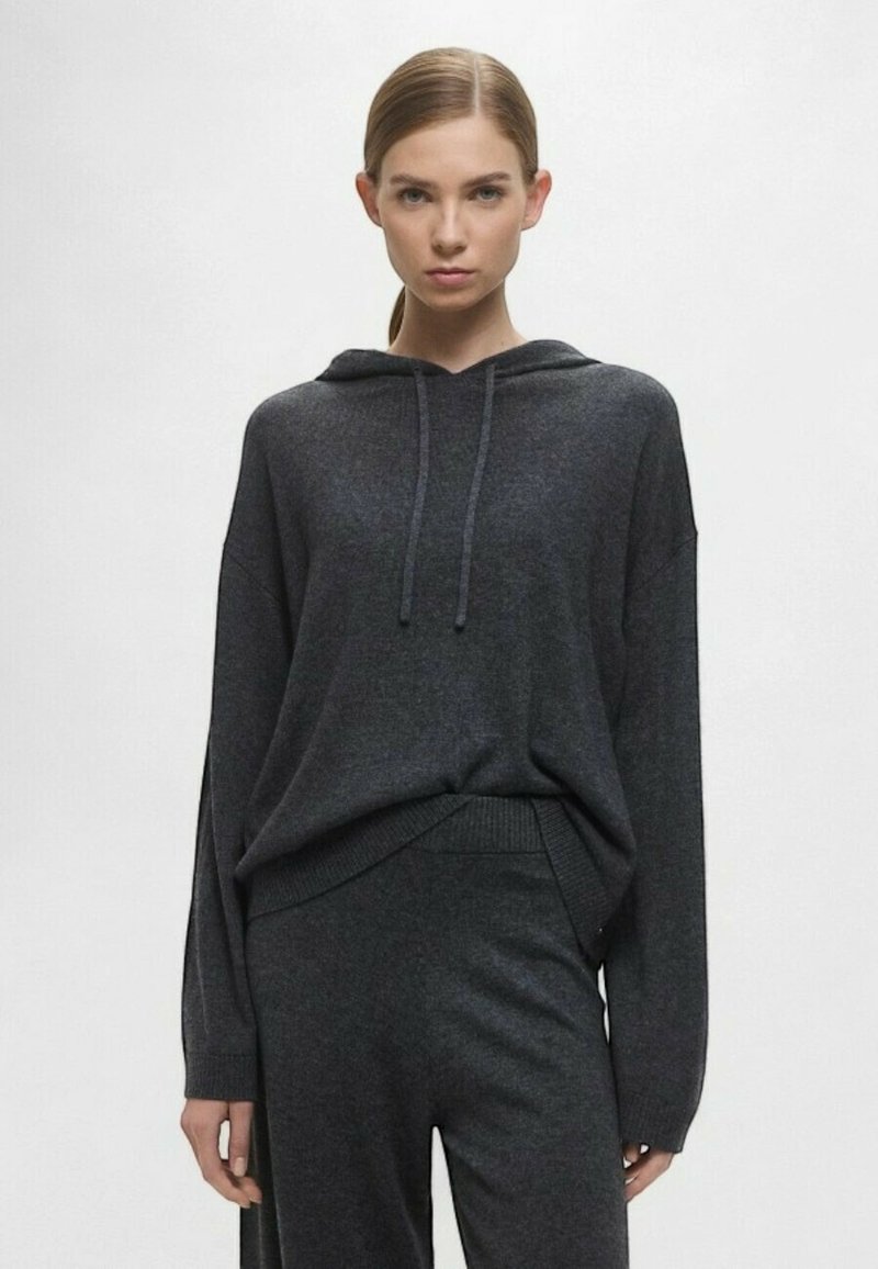 Felpa oversize grigia scura con cappuccio e coulisse, maniche lunghe, orlo a costine e texture morbida, abbinata a pantaloni coordinati.