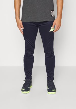 Granatowe sportowe spodnie wykonane z elastycznego materiału, o wąskim kroju, z prążkowanymi mankietami i małym zielonym logo na lewej udzie.