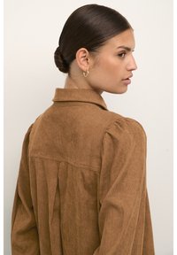 Veste chemise en velours côtelé brun avec col, manches longues et un détail de pli au dos. Texture lisse et coupe décontractée. Accents de couture subtils.