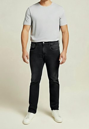 Mand stående iført en lysegrå kortærmet skjorte, sorte jeans og hvide sneakers på en neutral baggrund.