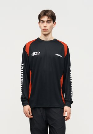 Mladić u crnoj sportskoj majici s dugim rukavima s crvenim panelima i bijelim logotipom te tekstom na rukavima, stoji na jednostavnoj svijetloj pozadini.