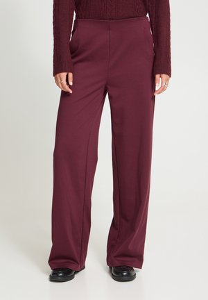 Femme portant un pantalon large bordeaux, un pull en maille assorti, des chaussures noires et des bagues en argent aux doigts.