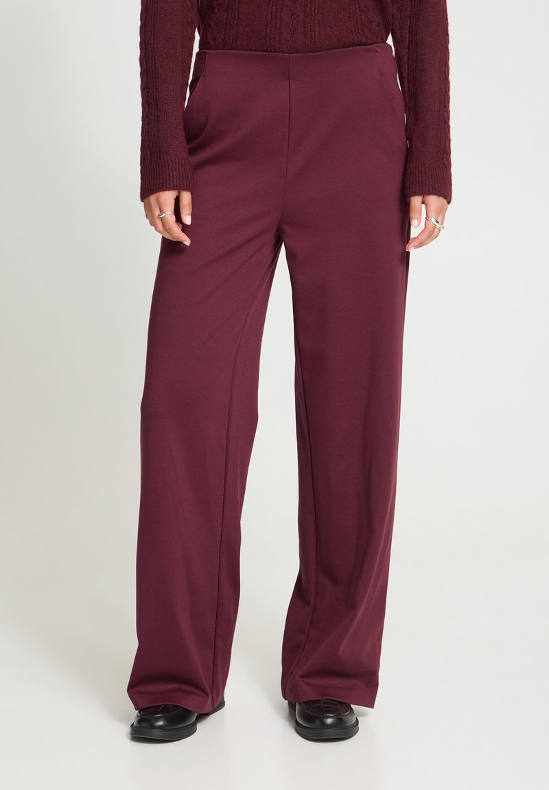 Femme portant un pantalon large bordeaux, un pull en maille assorti, des chaussures noires et des bagues en argent aux doigts.