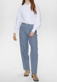 Vit långärmad skjorta med krage som kan knäppas; i kombination med blårandiga byxor; beige ankelstövlar; mot en ljus bakgrund.