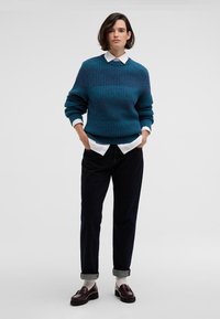 Τιρκουάζ πλεκτό πουλόβερ με υφαντό σχέδιο, λευκό πουκάμισο με γιακά από κάτω, σκούρες ρολαριστές παντελόνες και καφέ μοκασίνια. Απλή, καθημερινή εμφάνιση.