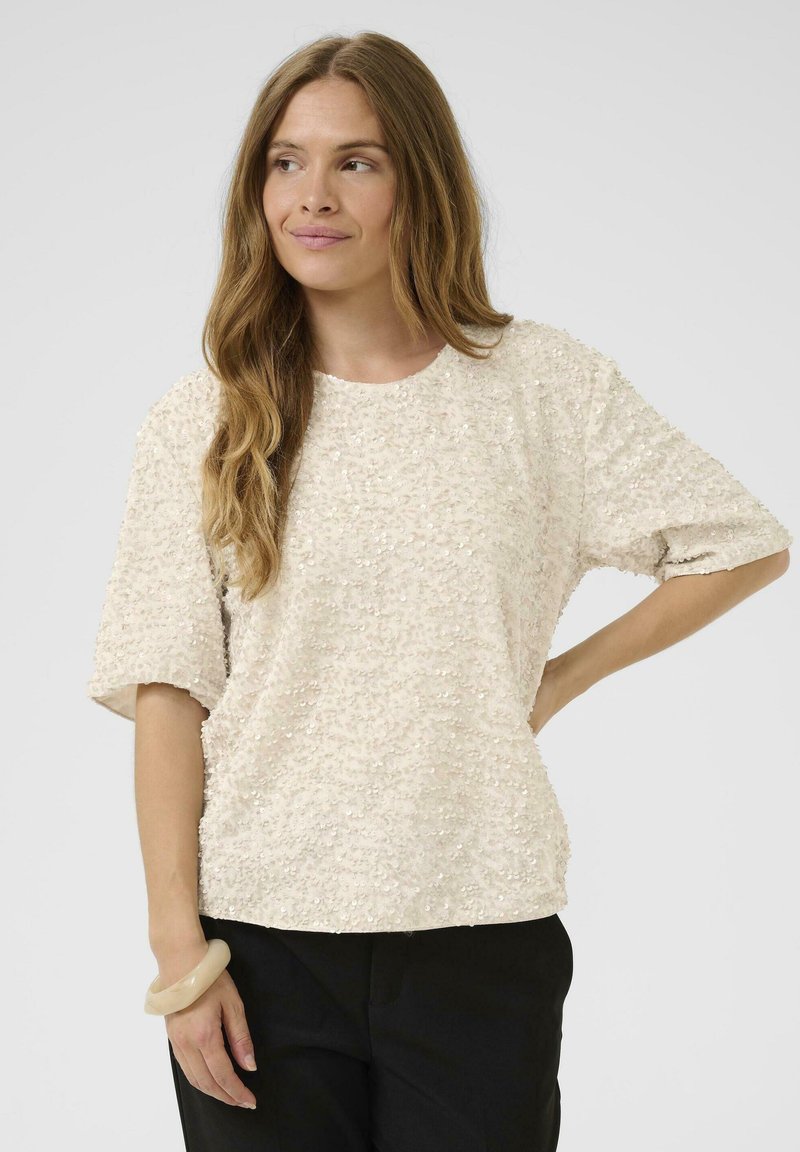 Beige krátký rukáv top s texturovanými, lesklými flitry. Hravý design, kulatý výstřih, v kombinaci s černými kalhotami a světlým náramkem na zápěstí.
