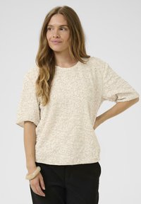 Beige īsām piedurknēm blūze ar teksturētām, spīdīgām sekvīnām. Kvadrāta forma, apaļa kakla izgriezums, savienota ar melnām biksēm un vieglu rokassprādzi uz rokas.