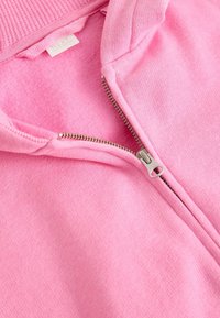 Lähikuva vaaleanpunaisesta fleecetakista, jossa on osittain avattu hopeinen vetoketju, näkyvissä resorikaulus ja pehmeä kangaspinta.