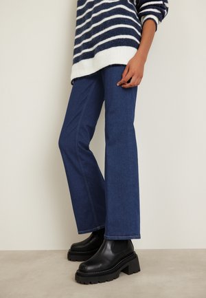 Henkilö, jolla on tummansiniset levenevät farkut, mustavalkoraidallinen villapaita ja mustat paksukorkoiset nilkkurit, seisoo sisätiloissa.