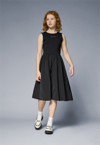 Juoda suknelė su prigludusia liemens dalimi ir plazdančiu sijonu. Ant krūtinės puikuojasi Calvin Klein logotipas. Dėvima su baltomis kojinėmis ir juodais loafers tipo batais su akcentais.