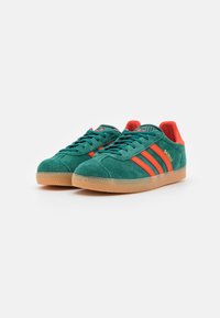 Green suede sneakers with red stripes, gum rubber sole, rounded toe, and padded collar. Features 'Gazelle' branding on the side.