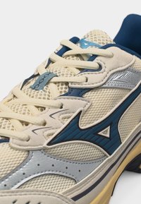 Beige og blå nett løpesko med kremhvite lisser, metallisk sølvdetaljer og en blå logo på siden.