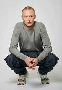 Pull gris à manches longues, pantalon à motifs foncés et baskets beiges. Gros plan sur des mains avec un tatouage. Le modèle est en position accroupie.