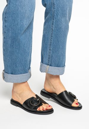 Schwarze Slide-Sandalen mit offenem Zehenbereich, versehen mit einem großen runden Metallakzent und einer glatten, lederähnlichen Oberfläche. Getragen zu hochgekrempelten blauen Jeans.