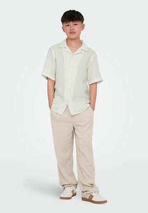 Jeune personne debout les mains dans les poches, portant une chemise légère à manches courtes, un pantalon beige et des baskets blanches avec des accents marron.