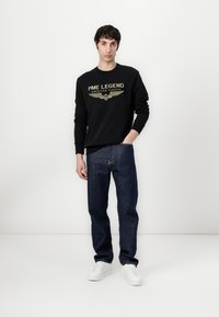 Felpa nera con la scritta "PME LEGEND AMERICAN CLASSIC" in lettere dorate, abbinata a jeans blu scuro e sneakers bianche su uno sfondo neutro.