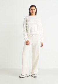Pulover alb tricotat cu un design din dantelă, asociat cu pantaloni largi crema și adidași gri. Detalii simple, cu texturi pe întreaga suprafață.
