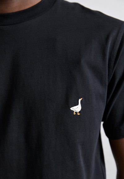 Mustvalge puuvillane T-särk, mille vasakus rinnapiirkonnas on väike bordüüritud valge hani oranžide jalgadega. Lühikesed varrukad ja ümar kaelus.