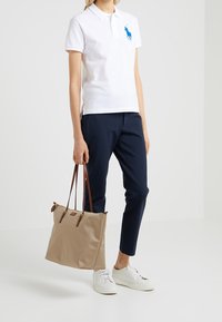 Vit polo shirt med blå logotyp, marinblå byxor, beige tote bag med bruna remmar och vita sneakers, mot en enfärgad bakgrund.