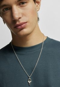 Silberne Kettenhalskette mit einem Vorhängeschloss-Anhänger, getragen über einem tealfarbenen Rundhalsausschnitt-T-Shirt. Nahaufnahme des Halsbereichs und Gesichts.