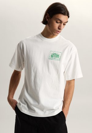 Mladý muž se středně dlouhými tmavými vlasy, oblečený v bílém tričku značky Vans se zeleným logem, ruce v kapsách, dívá se dolů, neutrální pozadí.