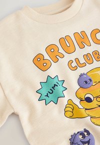 Kremowy sweter z kreskówkową stertą naleśników pokazującą kciuk w górę, jagodami z twarzami oraz napisami "BRUNCH CLUB" i "YUM!" w jasnych kolorach.