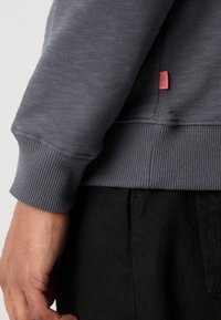 Sivi dukserica s rebrastim manžetama i rubom, s malom crvenom etiketom sa strane. Prikazuje teksturiranu tkaninu i kratak prikaz rukava.