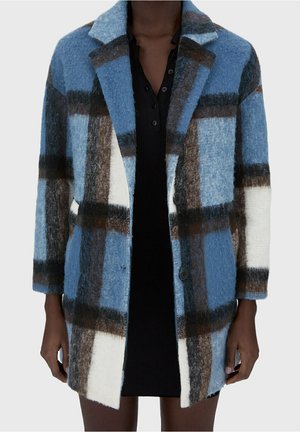 Mannequin portant un manteau mi-long à motif à carreaux bleu, blanc et marron sur une robe noire boutonnée.