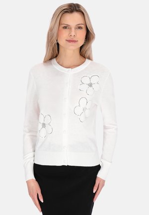 Nainen, joka pukeutunut valkoiseen napitettuun neuletakkiin, jossa on mustia strassikukkakuvioita, ja mustaan hameeseen, seisoo yksivärisen taustan edessä.