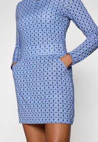 Blue patterned dress featuring a geometric design, long sleeves, a collar, and side pockets. Made of smooth fabric with a fitted silhouette.