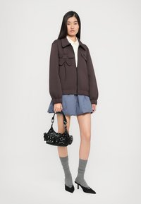 Chaqueta marrón con cremallera y detalle de lazo, camisa blanca con cuello, falda a cuadros azul, calcetines grises, bolso negro y zapatos de tacón negros.