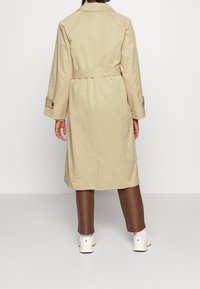 Beige trenchcoat met een tailleceintuur, lange mouwen en manchetten met knopen; gedragen over bruine leren broeken en witte sneakers.