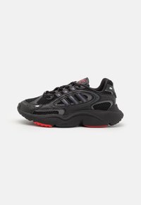 Black athletic shoe with mesh fabric, rubber sole, and red accents. Features padded collar, laces, and three stripe design on the side.