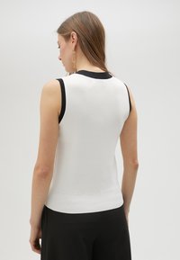 Bez rukava, bijeli top s crnim detaljima oko izreza. Mekana tekstura s pripijenim siluetom i rebrastim rubom. Minimalistički dizajn.