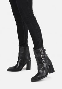 Botas de cuero negras hasta el tobillo con tacón cuadrado, punta afilada y múltiples detalles de hebilla. Textura suave con cierre de cremallera lateral.
