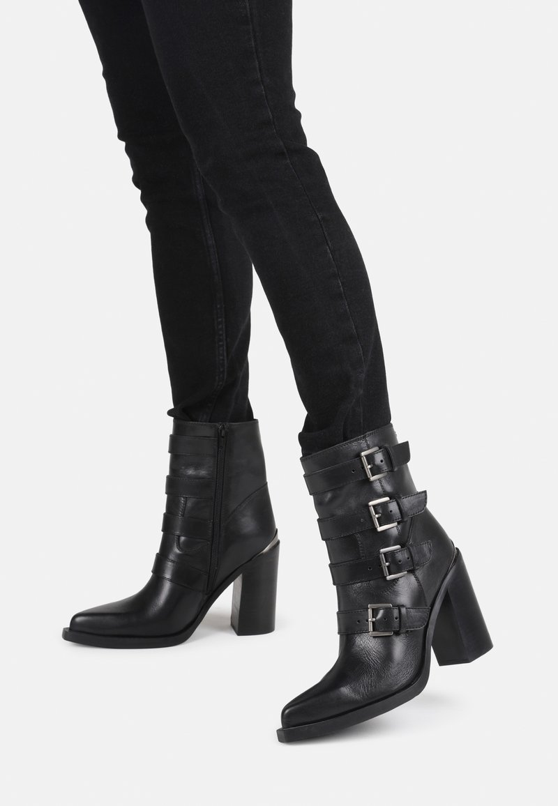 Botas de cuero negras hasta el tobillo con tacón cuadrado, punta afilada y múltiples detalles de hebilla. Textura suave con cierre de cremallera lateral.