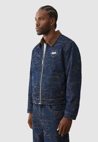 Spijkerjasje met bruin corduroy kraag, voorzien van een lichtbruin abstract patroon en een logopatch op de linkerborst. Ritssluiting.