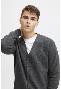 Cárdigan de lana gris con un profundo escote en V, cinco botones y puños acanalados. Lleva puesto sobre una camisa blanca, con una superficie suave y texturada.