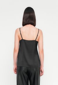 Femme aux cheveux bruns mi-longs portant un haut noir sans manches et un pantalon en cuir noir, se tenant devant un fond blanc uni.