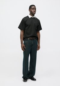 Black short-sleeve shirt with a white collar, single chest pocket; paired with loose dark denim jeans and black shoes, standing pose.