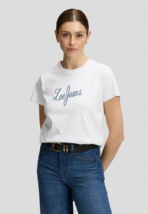 Frau steht nach vorne gerichtet, trägt ein weißes "Lee Jeans" T-Shirt, blaue Jeans mit schwarzem Gürtel, Haare nach hinten gebunden, neutraler Hintergrund.