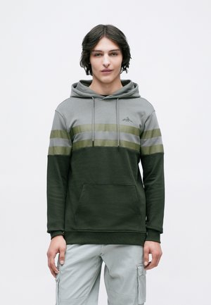 Jeune homme portant un hoodie coloré en vert et gris ainsi que des pantalons cargo gris clair, se tenant devant un fond blanc uni.